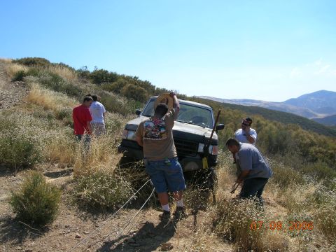1985 Ford Bronco - People work to free a stuck SUV on a remote, hilly terrain under a clear blue sky.