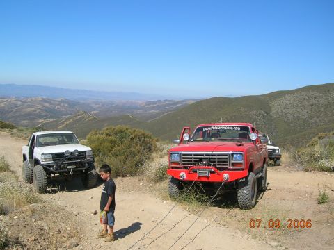 1985 Ford Bronco - Two off-road trucks are parked on a dusty trail with a scenic mountain view.