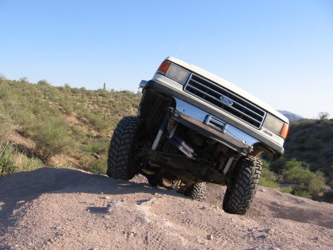 1990 Ford Bronco - A lifted Ford truck navigates a dirt hill with a desert landscape in the background.