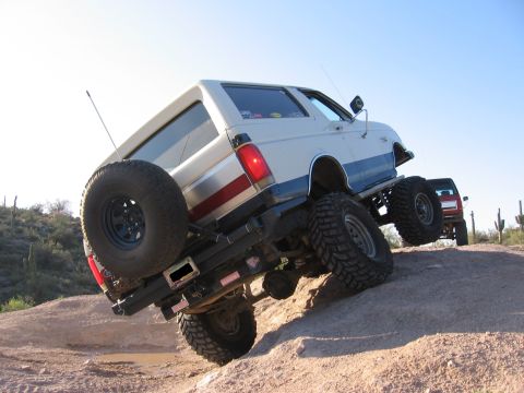 1990 Ford Bronco - A lifted Ford Bronco navigates a rocky desert terrain, with another vehicle visible in the background.