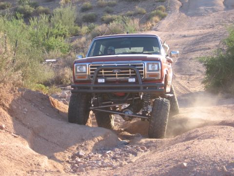 1990 Ford Bronco - This lifted Ford Bronco is navigating a challenging off-road trail with dust kicking up.
