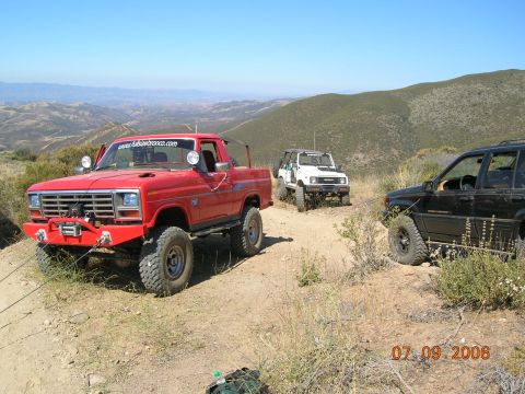 1985 Ford Bronco - Three off-road vehicles navigate a dusty trail in a hilly, arid landscape.
