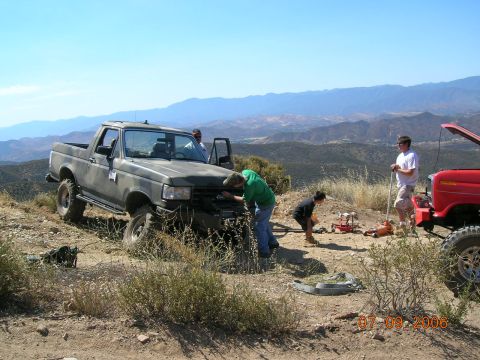 1985 Ford Bronco - Several people work on a distressed olive green Ford F-Series truck on a remote, mountainous terrain.