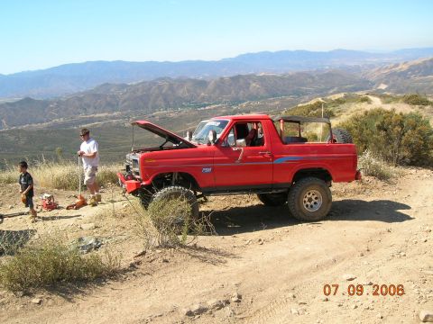 1985 Ford Bronco - Red Ford Bronco on a dirt trail, hood open, with two people standing next to it. Mountains in the distance.