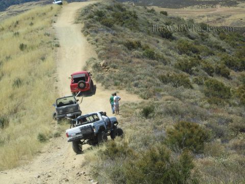 1990 Ford Bronco - red Jeep, a small truck and a lifted white 4x4 line up along a narrow dirt ridge trail as two people stand nearby.