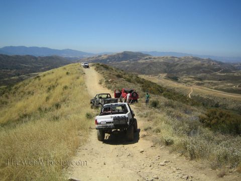 1990 Ford Bronco - lifted white open-top 4x4 leads vehicles along a dirt ridgeline trail with hazy mountains stretching to the horizon.