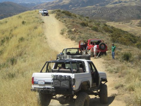 1990 Ford Bronco - white Ford open-top 4x4 follows a small truck and red Jeep along a dirt trail across grassy mountain ridges.