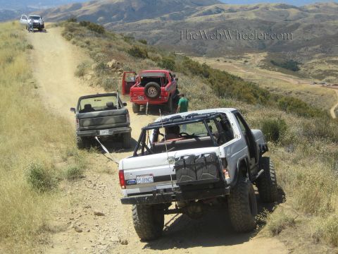 1990 Ford Bronco - white Ford 4x4, dark truck and red Jeep descend a winding dirt ridge trail as people walk between them.