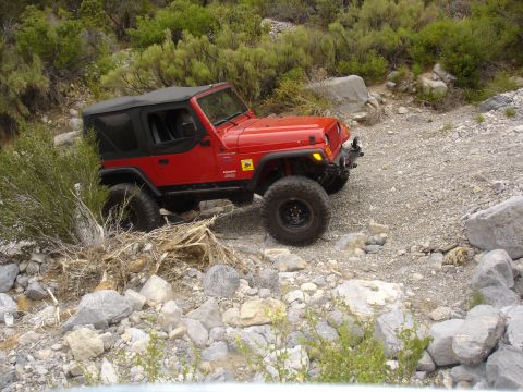 1996 Ford Bronco - Red Jeep Wrangler navigating a rough, rocky trail with sparse vegetation.