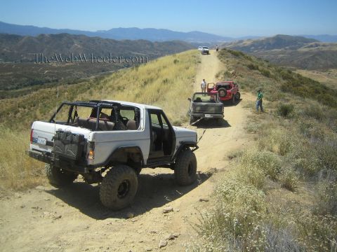 1990 Ford Bronco - white open-top 4x4 with a roll cage sits on a dirt ridge trail near a small truck, red Jeep and a few people.