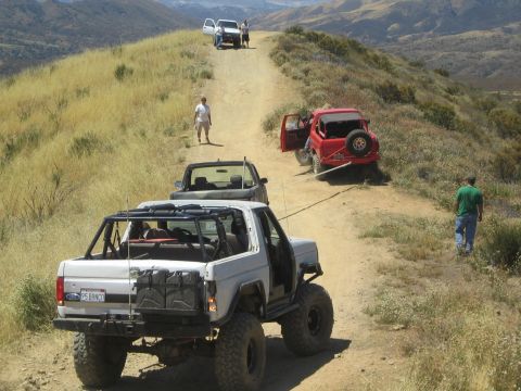 1990 Ford Bronco - white Ford 4x4 with a roll cage waits on a dirt hillside trail as a red Jeep and others gather farther up the slope.