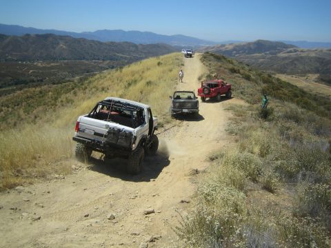 1990 Ford Bronco - white open-top 4x4 kicks up dust climbing a dirt ridge trail toward a small truck, red Jeep and waiting people.