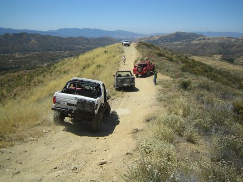 1990 Ford Bronco - Off-road trucks, including a silver pickup and a red rig, stage along a narrow dirt ridge trail in dry hill country.