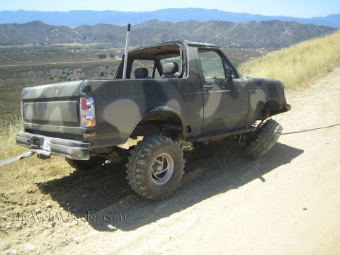 1990 Ford Bronco - dark camo-painted full-size Ford Bronco with its top removed sits lifted on mud-terrain tires on a dirt hillside.