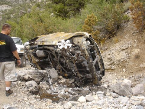 1996 Ford Bronco - A severely damaged and burnt vehicle lies on its side amidst rocks, with another SUV visible in the background.