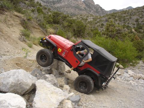 1996 Ford Bronco - Driver navigates a red Jeep Wrangler over a rocky obstacle on an off-road trail.