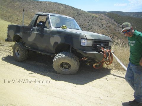 1990 Ford Bronco - man in a green shirt and cap stands beside a camo-painted lifted Ford Bronco rigged with a winch line on a hill trail.