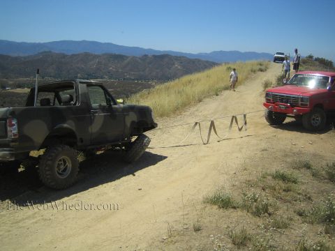 1990 Ford Bronco - camo-painted topless Bronco faces a red lifted truck across a dirt ridge trail as people stand nearby.