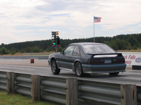 1989 Ford Mustang - A gray two-tone Fox-body Ford Mustang GT hatchback creeps toward the starting-line tree at a drag strip, shown from the rear three-quarter…