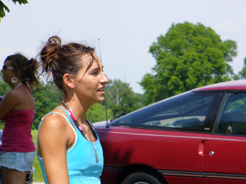 Woman in blue tank top next to a red car. Another woman in pink top is in the background