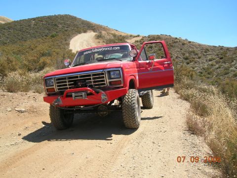 1985 Ford Bronco - A red truck is parked on a dusty trail with the driver's door open. The vehicle has been modified for off-road use.
