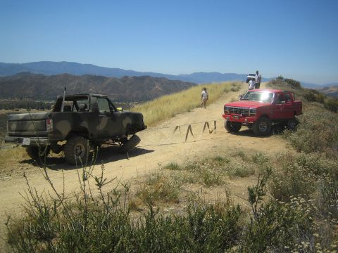 1990 Ford Bronco - camo topless Bronco and a red lifted truck sit on a dirt hilltop trail overlooking distant ranges.