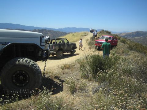 1990 Ford Bronco - lifted white Ford Bronco is rigged to tow a camo pickup along a narrow dirt ridge as bystanders watch.