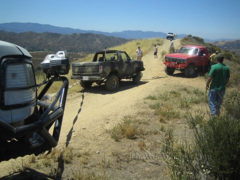 1990 Ford Bronco - white Bronco, a camo pickup and a red lifted truck line up on a dusty dirt ridge with people walking the trail.