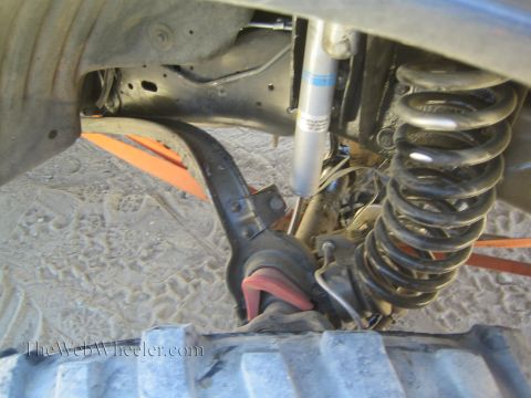 1990 Ford Bronco - Close-up beneath a lifted truck reveals a coil spring, shock absorber and orange recovery strap above the tire.