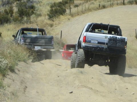 1990 Ford Bronco - white lifted Ford Bronco climbs a sandy wash on big tires alongside a camo pickup near a fence line.