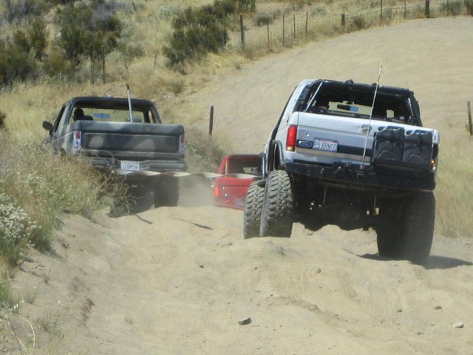 White lifted Ford Bronco climbing a sandy wash beside a camo pickup