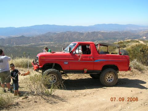 1985 Ford Bronco - Red Ford Bronco with lifted suspension parked on a dirt hill with mountains in the background.