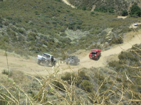 1990 Ford Bronco - Three off-road trucks, including a red rig, raise dust along a winding trail through a green scrub-covered canyon.