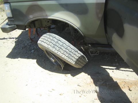1990 Ford Bronco - truck's rear tire lies folded sideways under the body after an apparent axle or suspension failure on a dirt trail.