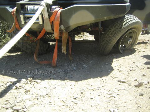 1990 Ford Bronco - front of an off-road truck shows a winch, frayed orange straps and a black-rim tire on broken rocky ground.