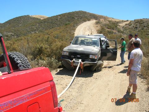 1985 Ford Bronco - A camouflaged Ford F-Series truck is stuck on a dirt road and is being recovered by another vehicle.