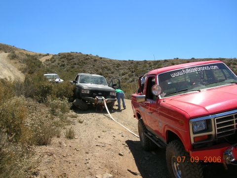 1985 Ford Bronco - A red Ford Bronco is parked on a dirt road, assisting a stuck black Ford Bronco in the bushes.