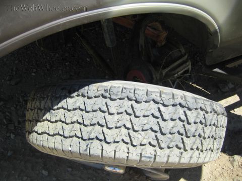 1990 Ford Bronco - worn all-terrain tire lies flat beneath a truck's rear fender, exposing the frame and suspension behind it.