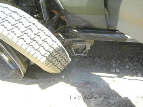 1990 Ford Bronco - tire is folded under a truck beside a bent step bar and tow-hitch receiver on dusty ground.