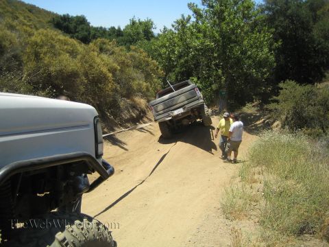 1990 Ford Bronco - lifted pickup tips on a steep dirt canyon trail as people stand nearby and another rig waits in the foreground.