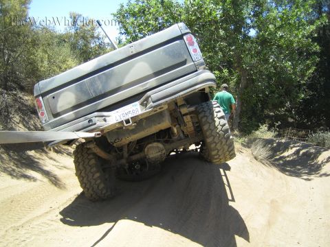 1990 Ford Bronco - rear of a lifted off-road truck climbs a steep sandy berm on oversized tires as a man in green stands beside it.
