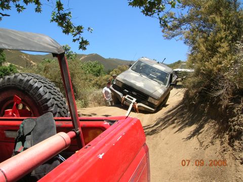 1985 Ford Bronco - A camouflaged pickup truck is stuck on a steep dirt path, being pulled by a red off-road vehicle.