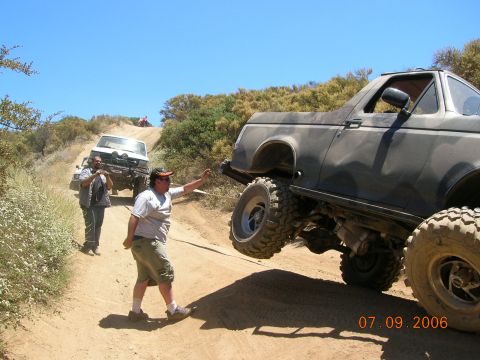 1985 Ford Bronco - A gray off-road SUV with a lifted suspension is being recovered on a dirt road, with another vehicle and people nearby.