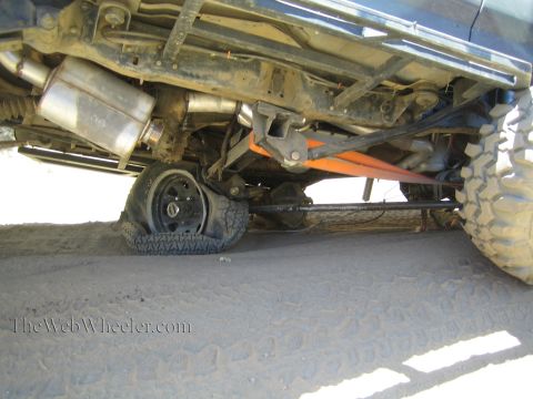 1990 Ford Bronco - View beneath a lifted truck reveals the catalytic converter, exhaust, driveshaft and axle with an orange strap over sandy ground.