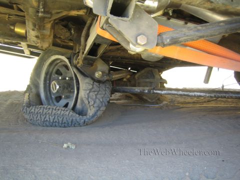 1990 Ford Bronco - folded tire on a black rim sits under a lifted truck's frame, with an orange strap and tie-rod visible on sandy ground.