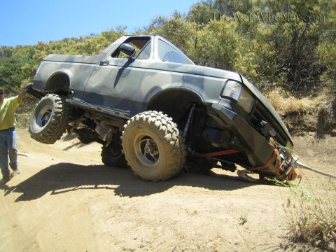 1990 Ford Bronco - camo-painted lifted truck balances at an angle on oversized tires on a dirt slope as a man in yellow stands at left.