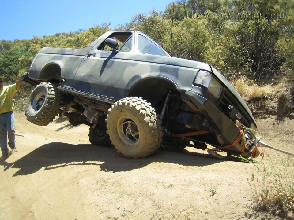 Camo lifted truck balanced on big tires on a dirt slope, man at left