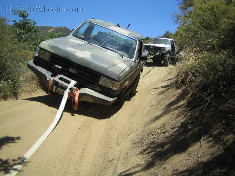 1990 Ford Bronco - camo-painted Ford pickup with a front winch sits on a rutted dirt off-road trail, recovery strap attached to its front.