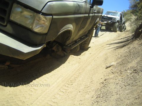 1990 Ford Bronco - Close-up of the front fender, headlight and off-road tire of a camo-painted Ford pickup on a dirt trail, another truck behind.
