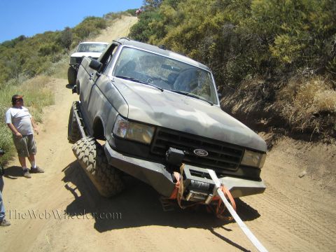 1990 Ford Bronco - camo Ford pickup with a front winch leans on a steep eroded dirt trail; a man stands beside it watching.